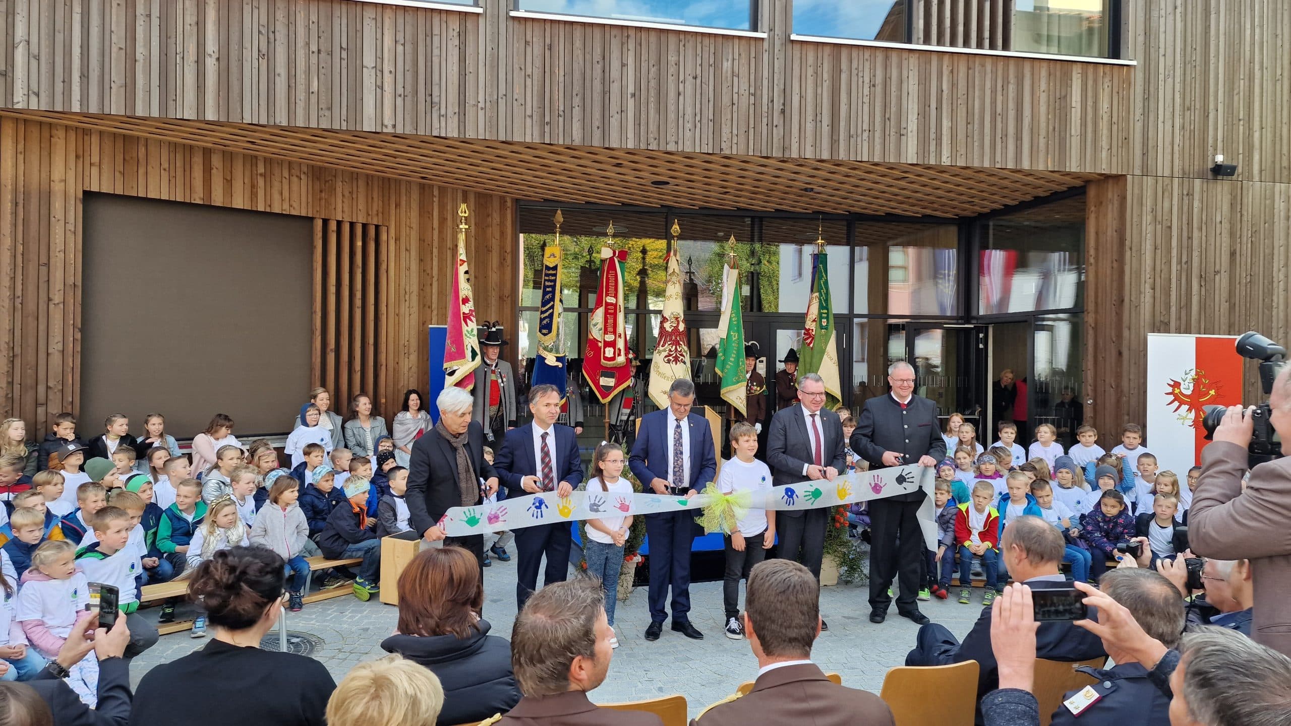 Eröffnungsfeier der Volksschule Breitenbach am Inn, mit Kindern und lokalen Würdenträgern, die gemeinsam ein Band durchschneiden.