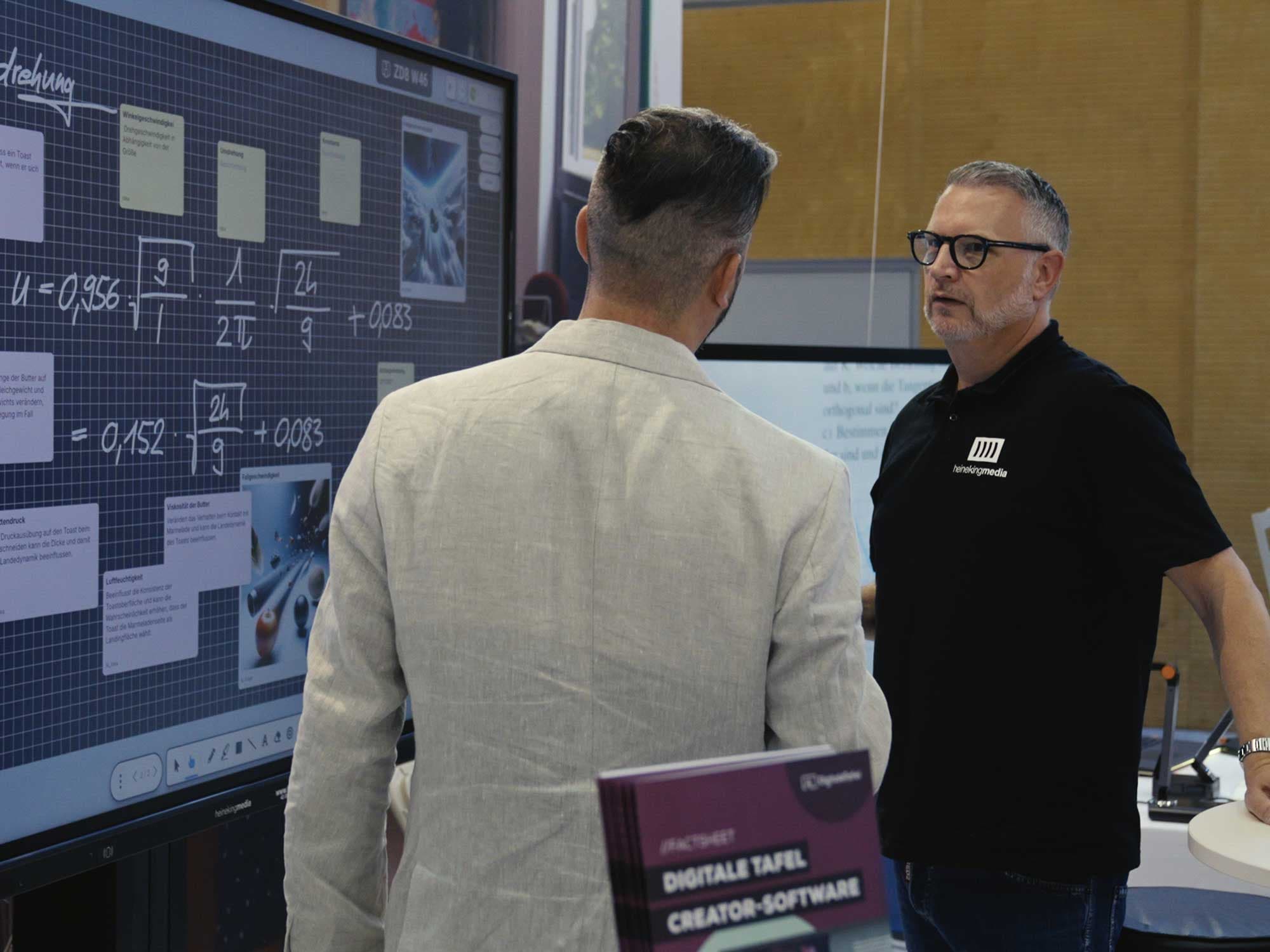 Wilfried Tollet von Heinekingmedia im Gespräch mit einem Besucher auf der Interpädagogica 2024 vor einem interaktiven Multitouch-Display.